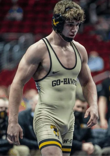 A muscular young man wearing a tight tan wrestling singlet and headgear stands in an arena.
