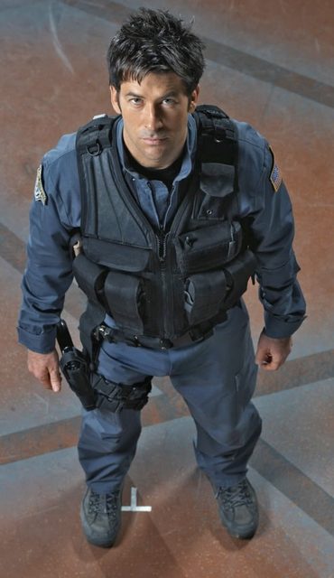 A masculine man in a blue tactical uniform with a black load-bearing vest and thigh holster looks up toward the camera.