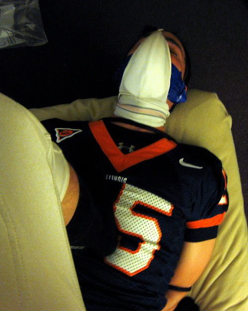 A man in a football jersey lies on his back with his hands bound behind him and an athletic sock pulled over his face as a makeshift hood.