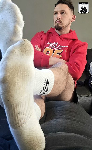 A bearded man in a red hoodie and shorts relaxes on a couch, extending his worn white crew socks toward the camera.