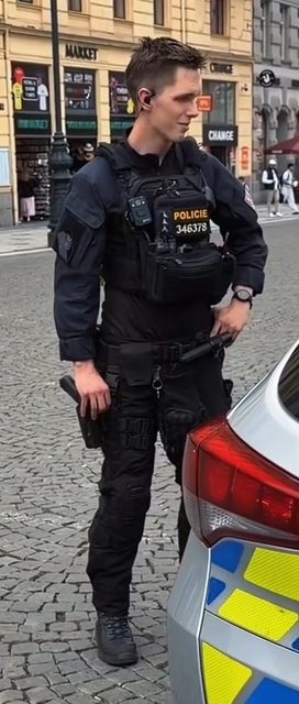 A young, fit man in a full black tactical police uniform with a gear-heavy vest and duty belt stands on a city street next to a patrol car.