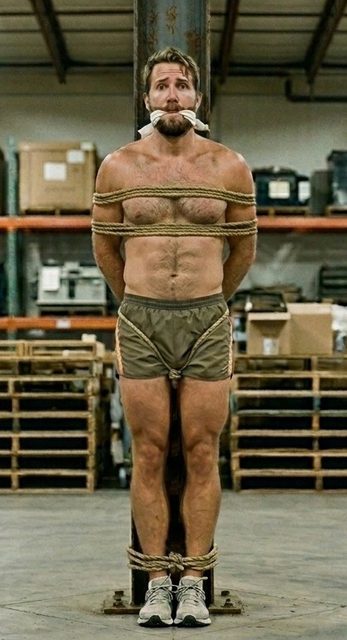 A muscular bearded man is tied to a warehouse support beam with rope around his chest, waist, and ankles while wearing a cloth gag.