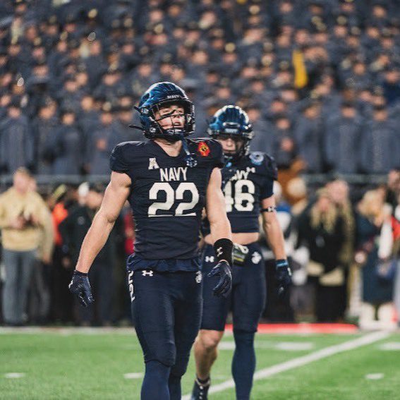 Two muscular athletes in full Navy football kits walk on the grass field in front of a stadium crowd.