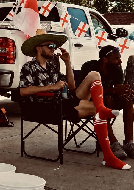 A man wearing long red Adidas football socks and black gym shorts sits in a folding chair beside a truck decorated with flags.