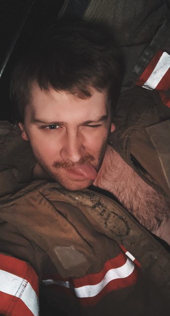 A hairy-chested man with a mustache and scruff wears a firefighter turnout jacket while winking and sticking his tongue out.