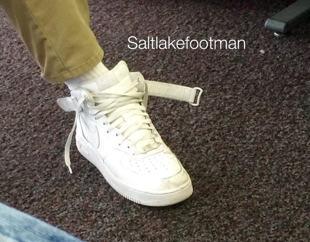 A close-up view of a foot wearing a white Nike Air Force 1 high-top sneaker with an undone ankle strap and white tube socks.