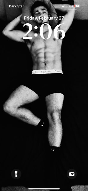 A muscular young man with defined abs and hairy legs lies on a bed wearing black Calvin Klein boxer briefs and Nike socks.