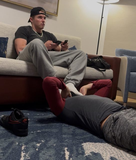 A muscular man relaxes on a couch while another man lies on the floor near his socked feet.