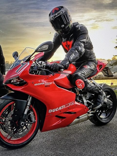 A person wearing full black leather motorcycle gear and a black helmet sits on a red Ducati sportbike outdoors.