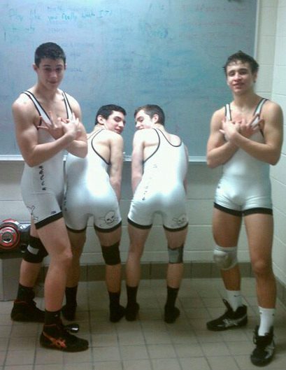 Four young men in white wrestling singlets and knee pads pose in a locker room, with two bending over to showcase their rear ends.