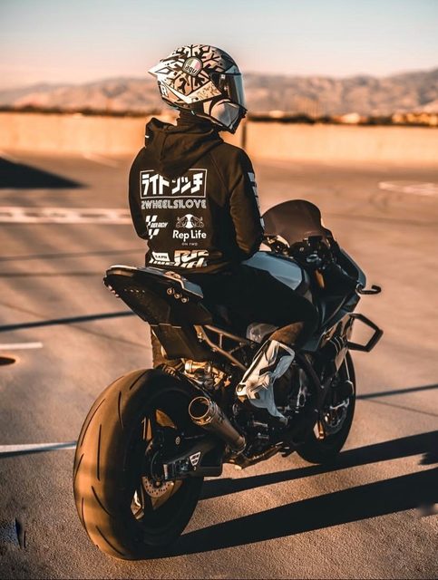 A person wearing a full-face racing helmet, a black hoodie, and white motorcycle boots sits on a sportbike in an outdoor parking lot at sunset.