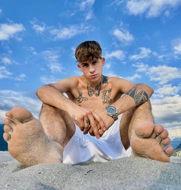 A tattooed young man sits on a sandy beach with his bare, sand-covered soles prominently displayed toward the camera.