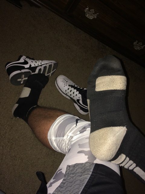 A man with hairy legs wears black and white athletic socks and patterned compression gear while sitting in a bedroom near his sneakers.