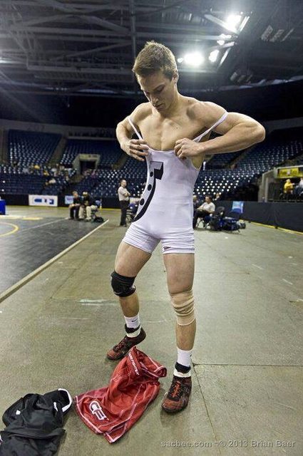 A muscular young man pulls on a white spandex wrestling singlet while standing in an indoor sports arena.