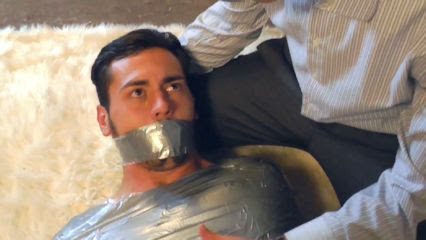 A young man bound and gagged with duct tape lies on a shag rug beside a man wearing a pinstripe dress shirt.