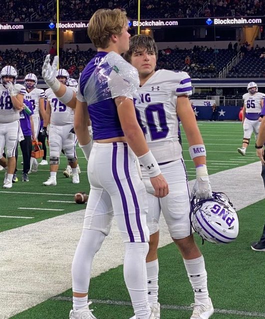 Two young athletic football players in tight purple and white uniforms converse on the field after a game.