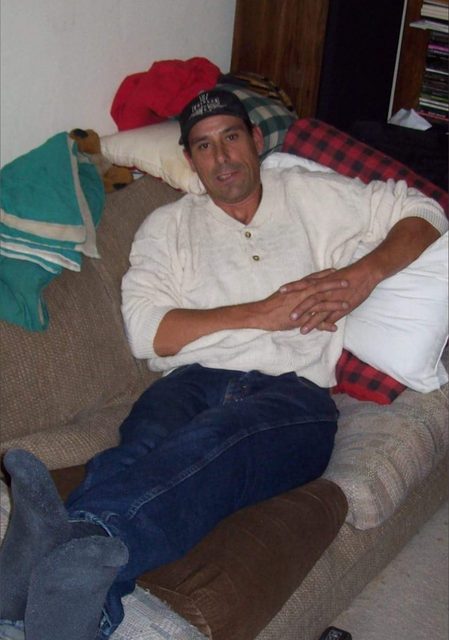A man lounges on a sofa wearing a shirt, jeans, socks, and a cap.