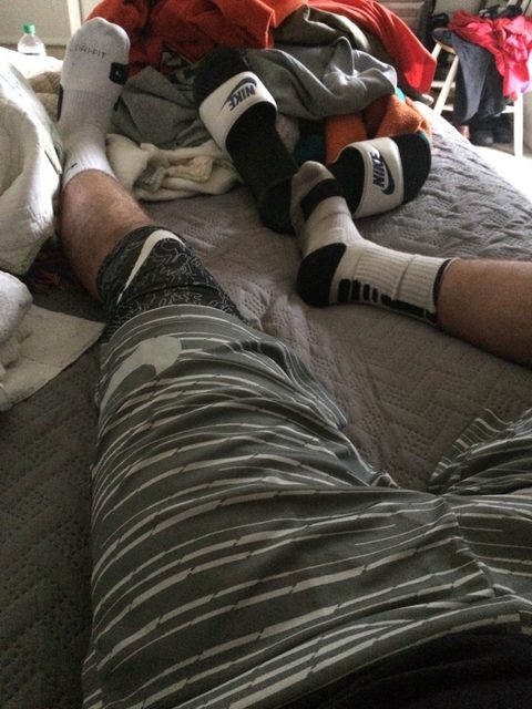 A man with hairy legs relaxes on a bed wearing athletic shorts, crew socks, and Nike slides.