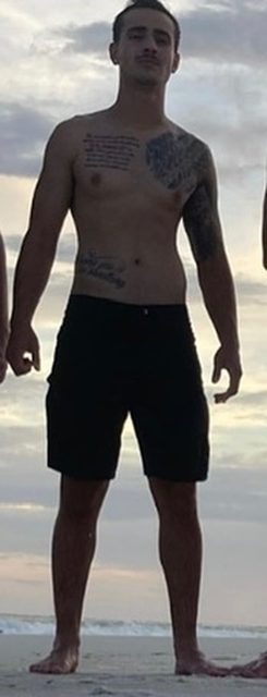 A shirtless, tattooed twunk poses on a sandy beach wearing black shorts.