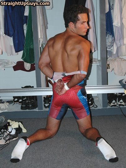 A muscular man kneels in a locker room with his hands bound behind his back, wearing a wrestling singlet pulled down to his waist.