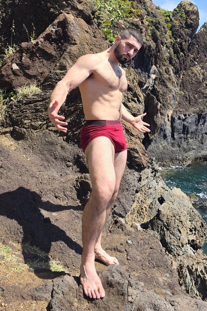 A bearded, athletic man wearing red underwear poses barefoot on a rocky coastal cliff.