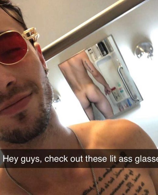 A bearded man takes a selfie wearing red sunglasses while his naked buttocks are visible in the bathroom mirror reflection.