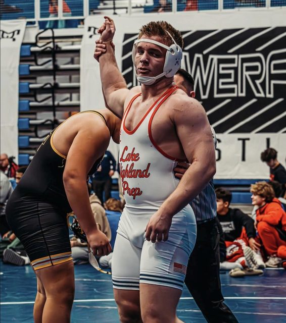 A muscular wrestler in a white and red spandex singlet has his arm raised in victory on the mat.