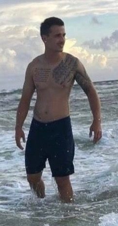 A shirtless young man with visible chest and arm tattoos stands in the surf at a beach.