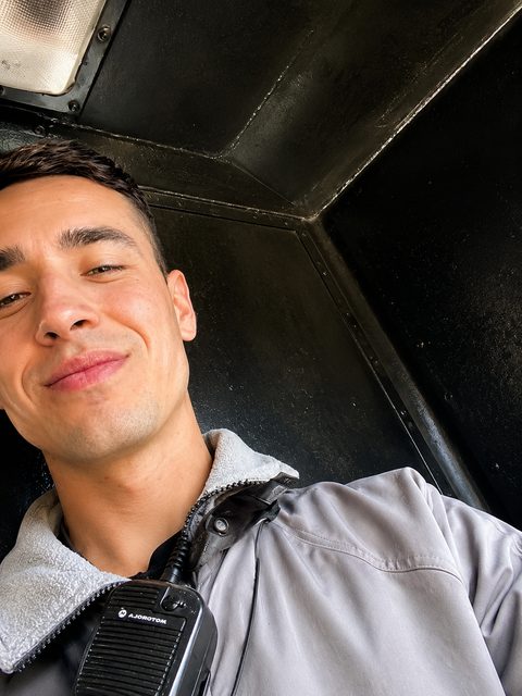 A smiling young man in a grey uniform jacket with a walkie-talkie clipped to his collar poses for a selfie in a dark, enclosed space.