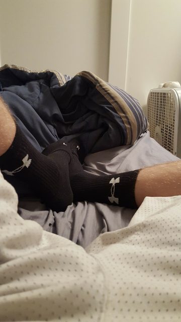 A POV shot of a man's hairy legs and feet wearing black Under Armour crew socks while lounging on a bed in mesh gym shorts.