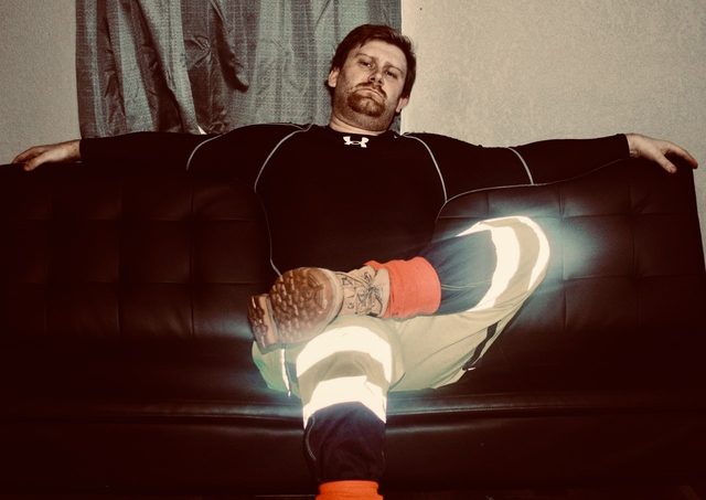 A bearded man in a compression shirt and high-visibility work trousers sits on a black sofa.