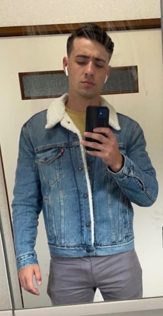 A young man with a buzzcut takes a mirror selfie while wearing a shearling-collared denim jacket and tight grey pants.