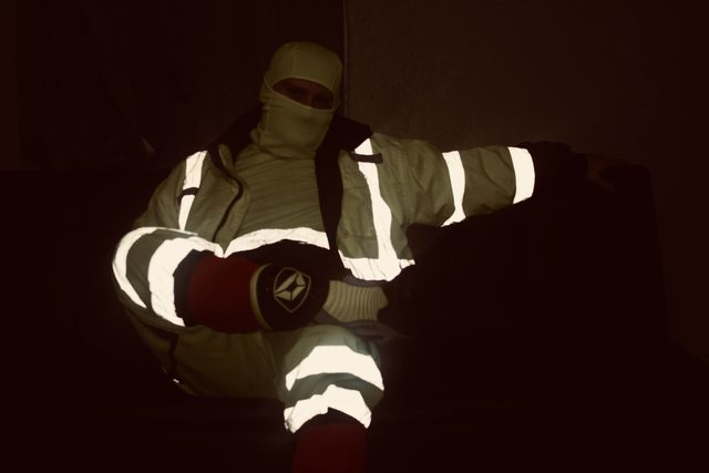 A man wearing high-visibility reflective workwear and a white balaclava sits in a dark room.