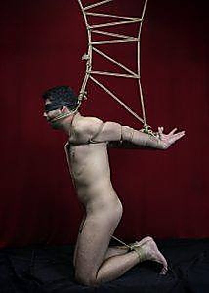 A nude man kneels blindfolded while bound in intricate rope shibari suspension bondage against a dark red backdrop.