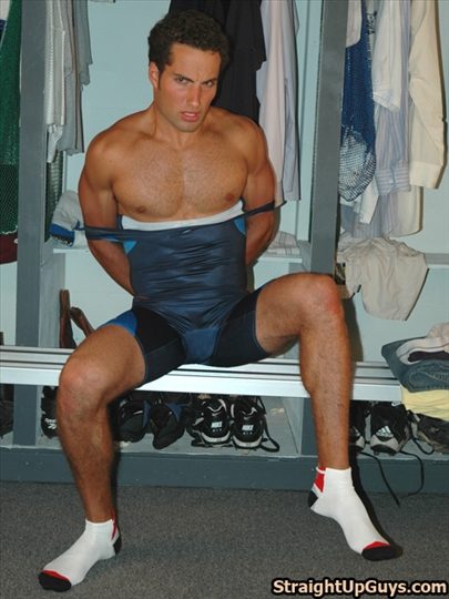 A muscular man in a wrestling singlet sits in a locker room with his arms restrained behind his back by the straps of his gear.