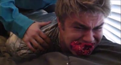 A man wrapped tightly in silver duct tape with a red cloth gag in his mouth lies prone on a bed while another person rests a hand on his back.