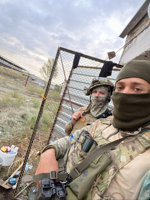 Two men in full military camouflage gear and tactical vests pose outdoors with balaclavas covering their faces.