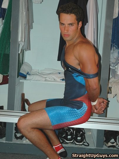 A muscular man in a tight blue and red wrestling singlet sits on a locker room bench with his hands held behind his back.