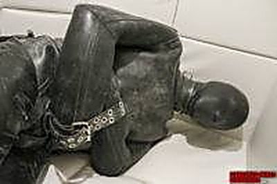 A person fully enclosed in a black leather suit and hood lies bound in leather straps on a white sofa.