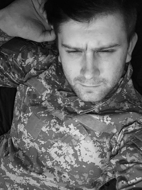 A black and white selfie of a scruffy man wearing a digital camouflage military uniform.