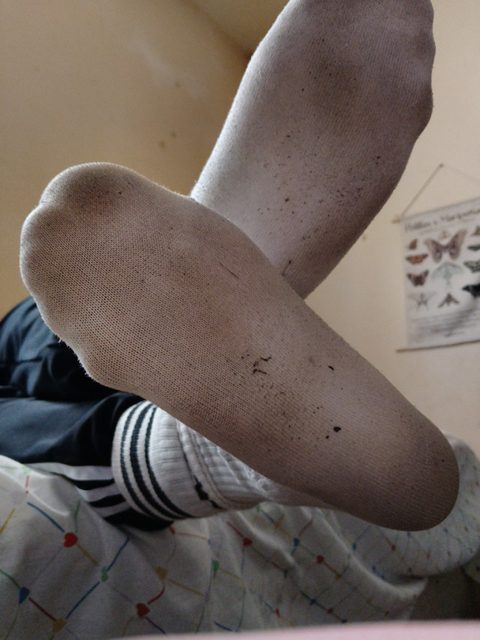 A man reclines on a bed, showcasing the soiled soles of his white crew socks paired with striped gym shorts.
