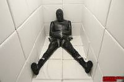 A person fully enclosed in a black latex gimp suit sits restrained on the floor of a white padded cell.