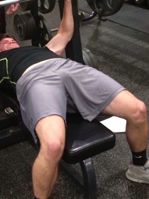 A muscular man in grey gym shorts lies on a weight bench, showing a visible print through his clothing.