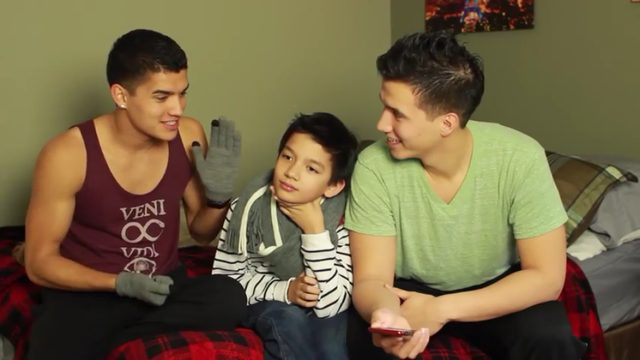 Two young men and a child sit together on a bed with plaid blankets in a bedroom.