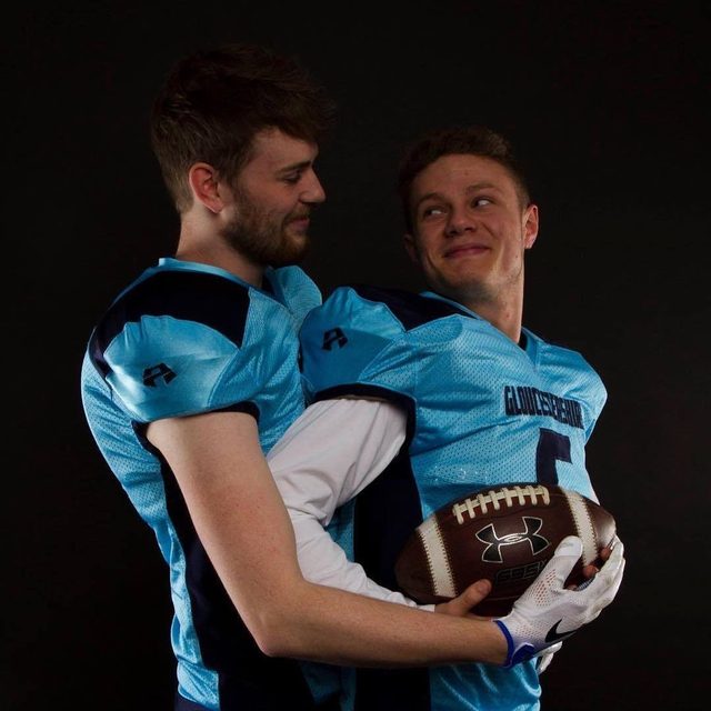 Two young men in light blue football uniforms embrace intimately while holding a football against a black studio background.
