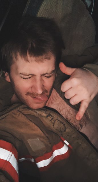 A man with a hairy chest wearing an open firefighter jacket makes a hand gesture with his tongue out.