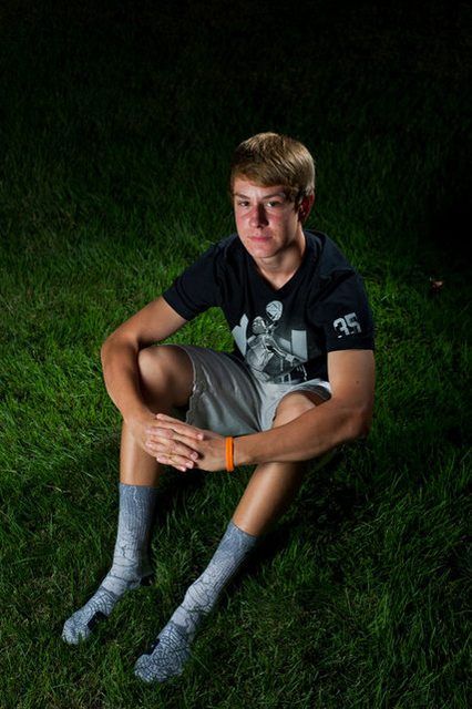 A young man sits on green grass at night wearing a black t-shirt, casual shorts, and graphic socks.