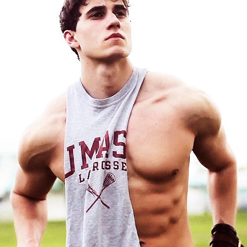 A muscular young man poses outdoors wearing a grey UMass lacrosse tank top pulled to one side to expose his defined chest and abdomen.