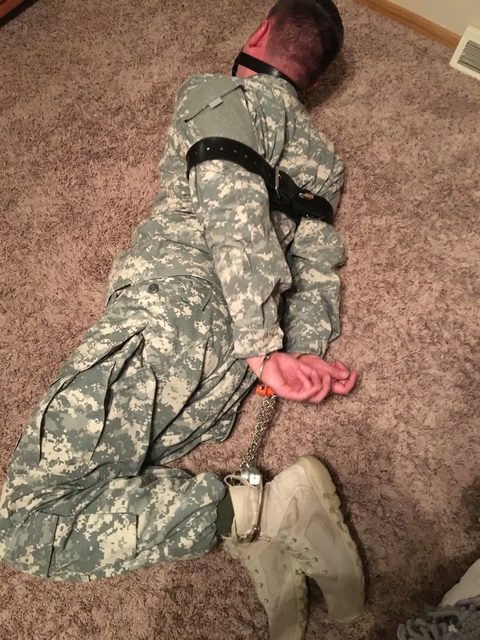 A man in a military camouflage uniform is restrained on a carpeted floor with a ball gag, handcuffs, and leather straps.