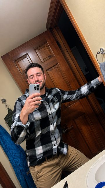 A bearded jock takes an amateur mirror selfie wearing a black and white plaid flannel shirt and tan pants.